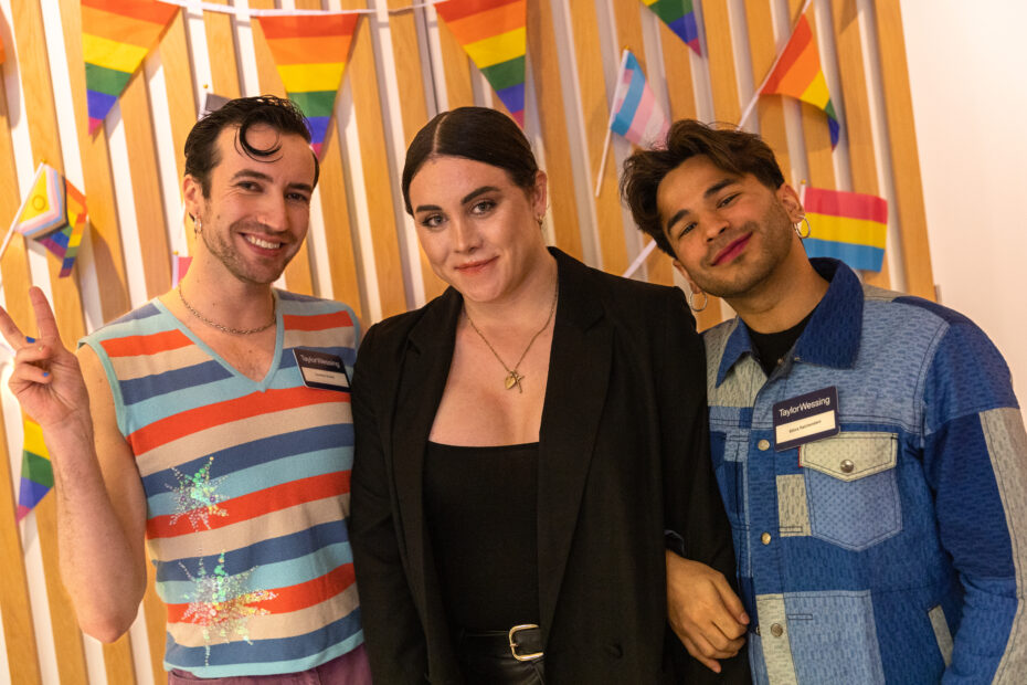 A photo of Dom, Charlie and Shiva standing in front of LGBTQ+ flags smiling. Shiva's arm is linked in with Charlie's and Dom is putting a peace sign up.