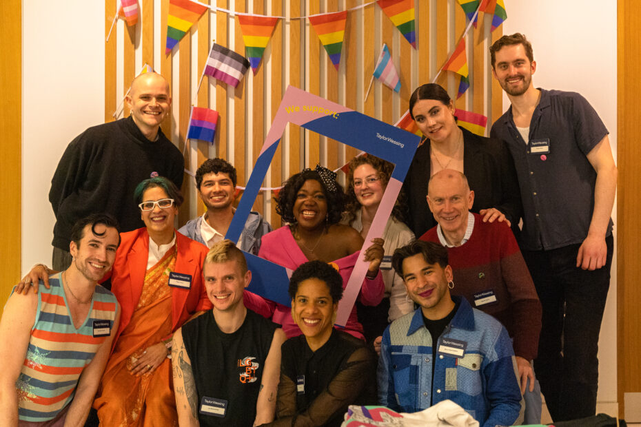 A group photo of the Centre team, Trustees and volunteers with new Patrons. Everyone is smiling standing behind a wall with LGBTQ+ flags.