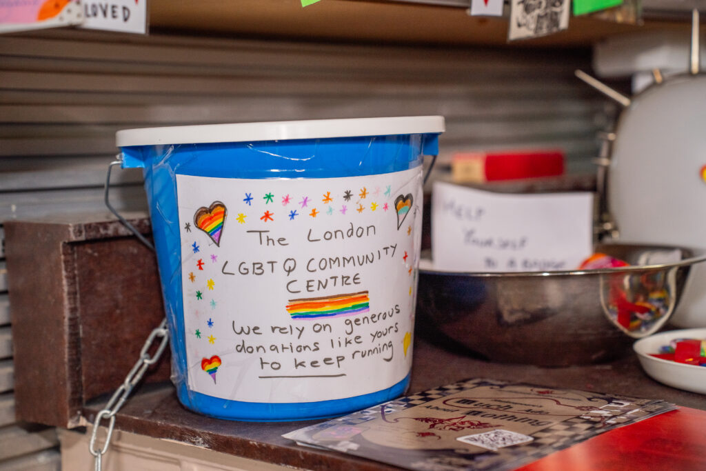 A blue donate bucket on the cafe counter. Handwritten message with stars and LGBTQ+ flags in hearts reads: 'The London LGBTQ+ Community Centre. We rely on generous donations like yours to keep running.'
